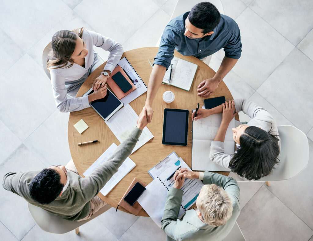Five people sitting around a round table, engaged in discussion with notebooks, tablets, and smartphones during their business English classes.