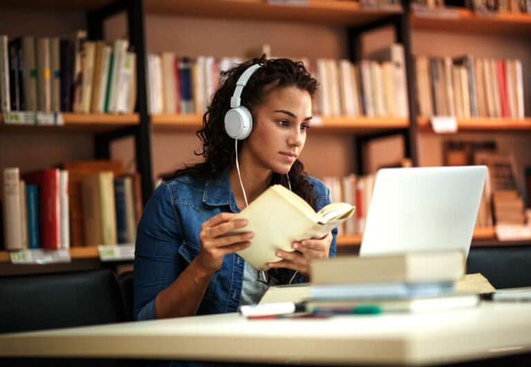 In a quiet library, a person wearing headphones reads a book while glancing at their laptop, immersing themselves in learning English online.