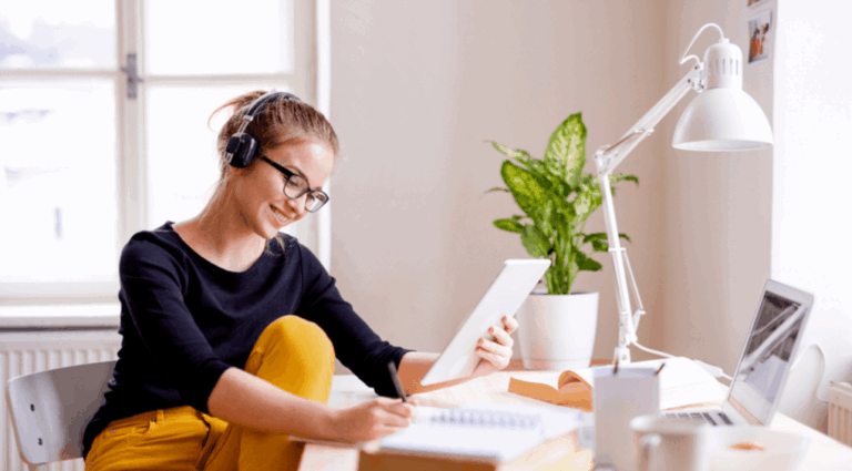A person engaged in online English learning sits at a desk with headphones on, using a tablet near a laptop and lamp. A potted plant adds a touch of nature by the window as they focus on their studies.