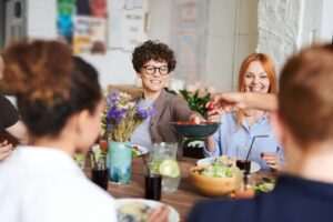 A group of people sit around a dining table, sharing a meal and lively conversation. They are practicing their language skills.