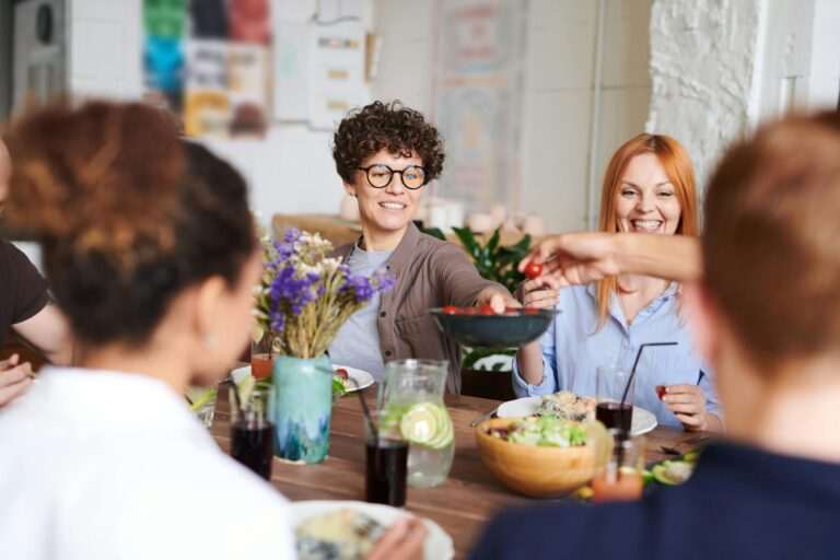 A group of people sit around a dining table, sharing a meal and lively conversation. They are practicing their language skills.