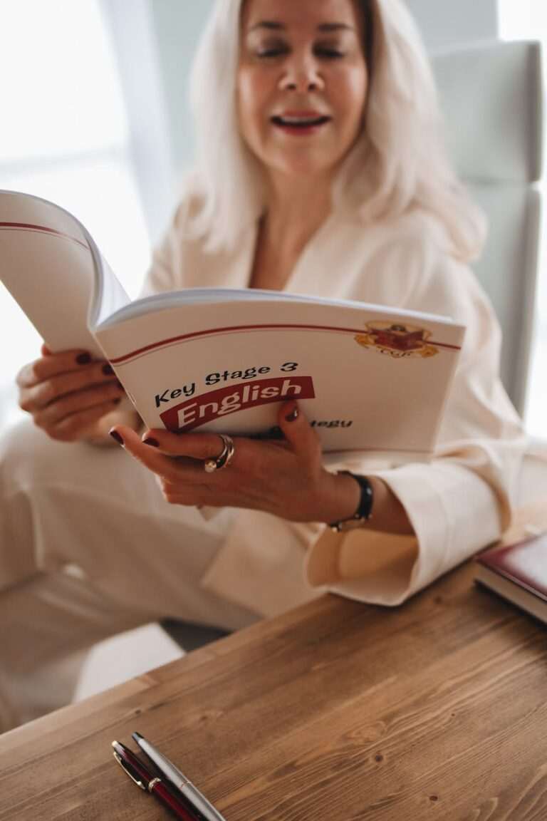 A woman in a white outfit reads a book titled "Key Stage 3 English" at a wooden table, focused on english practice exercises.