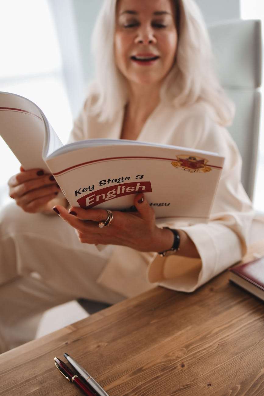 A woman in a white outfit reads a book titled "Key Stage 3 English" at a wooden table, focused on english practice exercises.